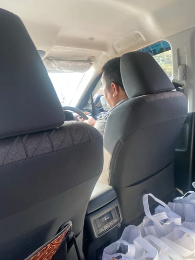 The image shows the interior of a car, viewed from the back seat. A man is visible in the driver's seat, seen from behind. The car appears to be parked or moving slowly in daylight, with sunlight streaming through the windows. The interior is mostly black and gray, with some white reusable shopping bags visible in the back. The environment outside the car is blurred but suggests a daytime setting, possibly a street or a parking lot. There is no specific information in the image to pinpoint a location in Manarap Lama, Indonesia.