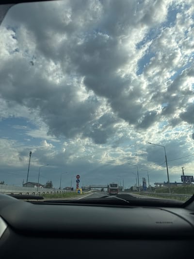 The image, taken from inside a vehicle, shows a view of the sky dominated by cumulus clouds. Through the vehicle's window, a roadside scene unfolds, featuring lampposts, road signs, and buildings. A red and white bus is visible in the distance. The location is identified as Усть-Сыны, Russia.