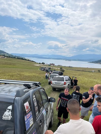 In the city of Rakitovo, Bulgaria, an outdoor gathering of off-road enthusiasts is taking place near the Batak Reservoir. The scene unfolds on a grassy hillside dotted with wildflowers, with the expansive reservoir and distant, hazy mountains forming the backdrop under a cloudy sky.

In the foreground, a dark-colored SUV, identifiable by its roof rack and numerous stickers on its rear window, is prominently positioned. These stickers include slogans like "Наско в колата" (Nasko in the car) and "OFF ROAD." A man in a white t-shirt is visible from the back, his head slightly turned.

To the right of this SUV, a group of men are conversing. One man in a black t-shirt with graphics and the word "OFFROAD" is holding a cigarette. Another man to his left wears a black t-shirt with Bulgarian text. Nearby, a woman with braided hair in a black t-shirt and shorts stands next to an open car door.

Further back, a line of SUVs and other vehicles is parked along a dirt road. Some vehicles have their doors open, and people are gathered around them, suggesting a break or preparation for an event. The vehicles appear to be prepared for off-road use, with their robust designs. In the distance, several buildings and a church spire can be seen on the shoreline of the reservoir. The overall impression is one of an organized off-road excursion or rally.