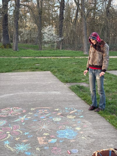 The image depicts an outdoor scene in the city of Saint-Mandé, France. A young individual stands on a light gray concrete path, positioned towards the right of the frame. They are wearing a brown faux fur hooded jacket with a red and blue plaid lining visible in the hood, a red shirt underneath, and distressed blue jeans with visible rips. The person holds a piece of white chalk in each hand, resting their hands at their sides, and directs their gaze downwards.
The concrete path in the foreground and midground is extensively covered with various colorful chalk drawings, including multiple floral patterns, stars, a butterfly, and abstract shapes, rendered in shades of pink, light blue, yellow, orange, and green. A single pink chalk stick and a small, clear plastic lid are present on the pavement near the bottom right corner of the image. A brown object, possibly a bag, is partially visible at the extreme bottom right.
The path is flanked by patches of vibrant green grass. In the background, a dense collection of deciduous trees with bare branches is visible, suggesting a late winter or early spring season. Among these trees, two smaller trees or bushes are in bloom, displaying white blossoms. A clear, light blue sky is visible above the tree line.
