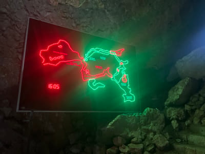An illuminated rectangular map display is centrally positioned within a dimly lit cave environment, specifically identified as Resavska Cave in Despotovac Municipality, Serbia. The display, approximately 1.5 meters in width, presents a detailed diagram of a cave system using embedded red and green LED lights to delineate pathways and halls. The left portion of the map is illuminated in red, labeling areas such as "УЛАЗНА ДВОРАНА" (Entrance Hall) and "ДВОРАНА СРАСЛИХ СТУБОВА" (Hall of Fused Pillars). The right portion is illuminated in green, indicating features like "КЕПИНА ДВОРАНА" (Kepa's Hall), "КОРАЛНИ КАНАЛИ" (Coral Channels), and "БОБАНОВА ДВОРАНА" (Boban's Hall). A red digital clock display on the lower left of the panel reads "16:05". The display panel is mounted on a metal stand. The surrounding environment consists of natural, rugged rock formations, rough cave walls, and scattered rock debris on the floor, all visible under the ambient dim lighting of the cave and the illumination emanating from the display itself. No individuals, direct actions, or interactions are present in the scene.