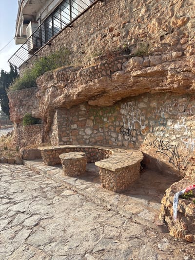 The image shows an outdoor stone seating area built into a rock face in Urbanización Cabo Roig, Spain. Three stone benches, each with a rounded seat and stone pillar supports, are arranged in a semi-circle on a paved stone walkway. The rock face behind the benches is textured and shows signs of weathering, with some graffiti visible on the stone wall. Vegetation grows in crevices of the rock face and along the top of the structure. To the left, a building with windows and a balcony is visible, supported by the rock face. The ground is paved with irregular, flat stones, and dry leaves are scattered on the surface. The lighting suggests it is daytime, with sunlight casting shadows. A roll of red and white striped tape is partially visible on the rocks to the right.