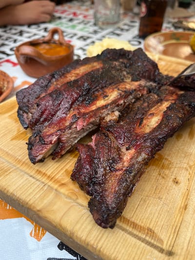 A close-up shot shows two pieces of heavily charred meat, likely ribs, resting on a light-colored wooden cutting board. The meat has a dark, almost black exterior with some visible red meat underneath. The cutting board is plain with visible wood grain. The setting appears to be a dining area, with blurry objects suggesting dishes and cutlery visible in the background. A tablecloth with a pattern of black and white geometric shapes is also present in the lower right corner.