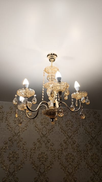 A close-up, low-angle shot shows a decorative chandelier hanging from a white ceiling. The chandelier has a gold-tone metal frame with multiple arms, each holding a frosted glass shade and a clear crystal pendant. Three LED light bulbs are illuminated in a warm white tone. The background is a textured wallpaper with a damask pattern in gold and brown hues. The image is taken indoors, likely in a residential setting within Занарацкі сельскі Савет, Belarus. There are no people or animals visible in the frame.