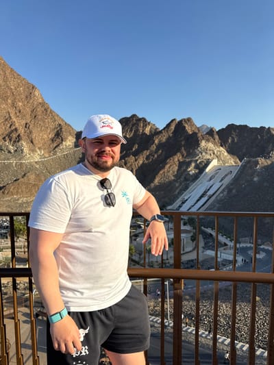 An adult male stands on an elevated viewing platform in Hatta, United Arab Emirates, overlooking the Hatta Dam and the surrounding Al Hajar Mountains. The man, with fair skin, short brown hair, and a beard, is positioned slightly left of center, looking directly at the camera with a subtle smile. He wears a white baseball cap featuring a red and black "Paul&Shark Yachting" logo, a white short-sleeved t-shirt with a light blue palm tree and crescent moon emblem on the left chest, and black shorts with a white design on the left leg. Black sunglasses are suspended from his t-shirt collar. A blue-grey smartwatch is visible on his left wrist, and a light teal fitness band is on his right wrist. His left forearm rests on a dark brown metal railing that spans horizontally across the lower foreground. The background reveals rugged, rocky mountains under a clear, cloudless blue sky. A large mural depicting two portraits, identifiable as Sheikh Zayed bin Sultan Al Nahyan and Sheikh Khalifa bin Zayed Al Nahyan, is prominently displayed on the face of the Hatta Dam to the right. Below the dam, a winding paved road with several individuals walking on it meanders through the landscape, alongside sparse desert vegetation and low-rise structures.