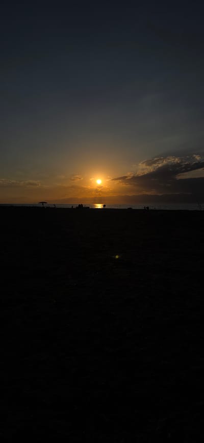 This is a vertical, full-frame photograph of a beach scene at sunset. The sun is low on the horizon, casting a golden glow and reflecting on the water. The sky transitions from a dark, deep blue at the top to a warm orange and yellow near the horizon. Clouds are scattered across the sky, some catching the sunlight.
In the middle ground, a silhouetted shoreline is visible, with faint mountain ranges in the distance. Along the beach, several small, dark figures are scattered, some appearing to be individuals or small groups of people. One figure is partially obscured by what could be a beach umbrella. The water's surface reflects the bright sun.
The foreground is almost entirely black, showing the dark sand of the beach. A faint, yellowish-green lens flare is visible in the lower middle section of the foreground. The overall mood is serene and tranquil, capturing the end of a day at the beach. The scene is located in Boz Beshik, Kyrgyzstan.
