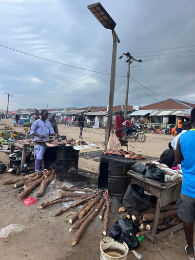 The image depicts an outdoor market scene in Oko Amakom, Nigeria. In the foreground, a man in a light purple traditional outfit is grilling meat on a makeshift barbecue, which consists of metal drums and a grate. Several pieces of raw meat are laid out on the grill. The fire beneath the grill is fueled by logs of wood. To the left of the grilling station, a pile of raw tubers, likely cassava, is visible. Various black plastic bags and some white plastic sheeting are scattered on the ground around the market stalls.
In the mid-ground, several other market stalls with corrugated metal roofs are visible, along with a few people engaged in market activities. A motorcycle with a rider and passenger is moving through the scene. In the background, more market stalls and structures extend into the distance. The sky is overcast with dramatic clouds. The overall impression is a bustling, everyday market scene in a Nigerian town.