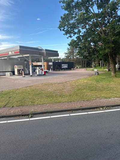 This image depicts a gas station with a convenience store, identified as "MOMENTS & MORE," located in Bladel, Netherlands. The scene is captured from a roadside perspective, showing a portion of the road with a white line. The gas station features multiple pumps under a covered canopy. A white truck is parked at one of the pumps, along with a car. The convenience store is a modern, dark-colored building with large windows. Two individuals are seen walking near the store. The environment is somewhat rural, with a grassy area and trees surrounding the gas station, suggesting it might be on the outskirts of town or along a highway. The sky is blue with some scattered clouds.