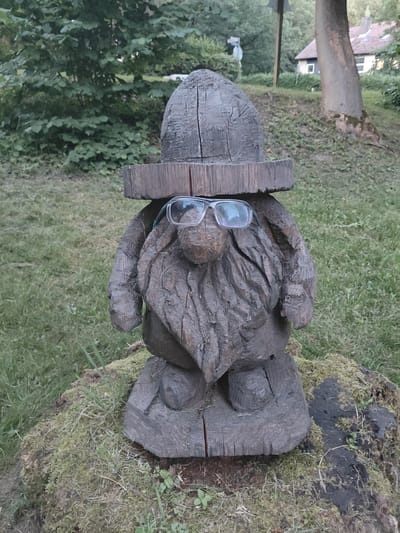 A weathered wooden gnome statue, complete with a hat and glasses, stands on a moss-covered tree stump in a grassy area. In the background, a house with a red roof and windows can be seen, along with trees and foliage. The overall scene suggests an outdoor setting, possibly a garden or park, in Bad Lauterberg im Harz, Germany.