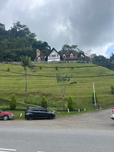In Ringlet, Malaysia, a building with a Tudor-style architectural design, featuring white half-timbered walls and a dark roof, is situated atop a verdant, grassy hill. The building has multiple windows with dark frames and a prominent brick chimney. A white fence runs along the crest of the hill in front of the building, and several flags, including the Malaysian flag, are visible. The hill is sparsely populated with small, conical evergreen trees, and two large palm trees with fan-shaped leaves stand prominently on the slope. Several power lines are strung across the hillside. At the base of the hill, a paved road runs horizontally across the foreground. Two cars are parked on the shoulder of the road: a red sedan on the left and a dark-colored hatchback on the right. The environment is characterized by a dense, green forest in the background, and the sky is overcast with thick, gray clouds.