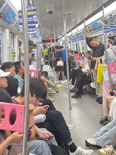 The image shows a crowded subway car in 龙溪街道, China. Passengers are sitting and standing, with many using their cell phones. The interior of the subway car has advertisements and safety warnings posted. Several passengers are leaning on the handrail.