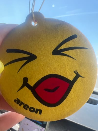 A yellow, circular Areon brand car air freshener, designed with a winking emoji face featuring a red tongue protruding, is centrally positioned and held by human fingers on the left. The air freshener has two stylized black eyes, one of which is closed in a wink, and the word "areon" printed in black lowercase letters at the bottom-left. A string for suspension is visible at the top, passing through a punched hole. The background reveals a clear blue sky with faint, high-altitude wisps, seen through what appears to be a vehicle windshield. Below the air freshener, the dark, textured interior of a vehicle's dashboard or console is visible, along with a partial view of a digital screen displaying a map or navigation interface on the lower left. The scene is located in Kolarovo, Bulgaria.