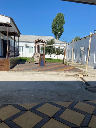 The image depicts an outdoor scene in what appears to be a communal area or a schoolyard in Karabudakhkent, Russia. The foreground is dominated by a cracked concrete surface, leading to a paved area with colorful, square tiles in shades of red, yellow, and green. Beyond the tiled area, a wooden children's play structure with a slide and ladder is visible.
The background features several low-rise buildings. To the left, a white building with a white, corrugated roof and a covered porch with a metal railing is partially visible. A sign with Russian text, possibly indicating an exit, is affixed to the wall. To the right, another white building with a corrugated roof and multiple windows is seen. Air conditioning units are mounted on the walls of these buildings. In the center of the frame, a tall, slender green tree stands against a clear blue sky. The overall environment suggests a sunny day, with shadows cast by the buildings and the play structure. No people are present in the image.