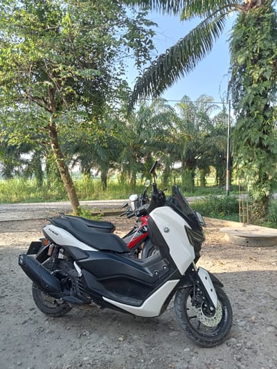 Two motorcycles are parked outdoors on a gravelly surface. The motorcycle in the foreground is white and black, with "NMAX" branding visible. The motorcycle behind it is red and black. The scene is set in a tropical environment with palm trees and lush green vegetation. The sky is clear and blue. In the background, a small concrete structure with a circular opening is visible. The lighting suggests it is daytime, likely morning or afternoon, given the shadows. The location is Meunasahrayeuk, Indonesia.