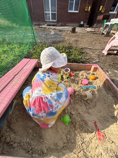 A child, wearing a white eyelet hat and a colorful, paint-splattered dress, lies on their side in a wooden sandbox filled with sand. Toys are scattered around the child and in the sand, including a blue plastic shovel, a red plastic shovel, a pink cup, an orange ball, and a green frog toy. The sandbox has pink wooden edges. To the left of the sandbox, a brick building with windows is visible, along with some green fencing and dry grass. A pink plastic slide is also partially visible in the background. The lighting suggests it is daytime, possibly late afternoon due to the shadows. The scene appears to be an outdoor play area.