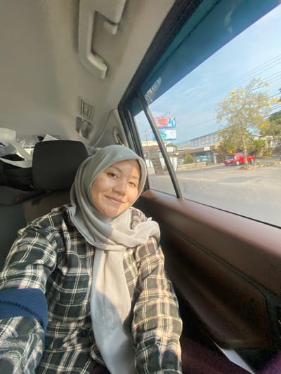 A woman wearing a light grey hijab and a plaid shirt is seated in the passenger seat of a car. She is looking at the camera and smiling. The interior of the car is visible, including the brown leather seat and the grey headliner. Through the window, a street scene in Mandar Sari, Indonesia, is visible. There are trees, buildings, power lines, and other cars on the road. The sun is shining, casting shadows on the scene.