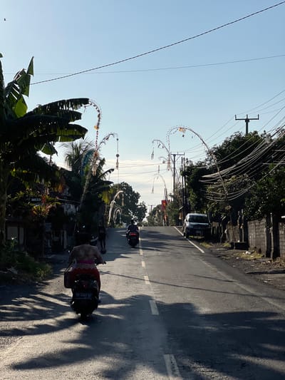 The image depicts a street scene in the city of Kubu, Indonesia, bathed in the warm light of what appears to be early morning or late afternoon. The road, asphalted and marked with a white line down the center and a dashed white line on the right edge, slopes gently upwards. On the left side of the road, there are buildings with visible roofs, and lush green banana trees with broad leaves casting long shadows across the pavement. Traditional Balinese decorations, known as "penjor," adorn poles along the roadside, featuring curved bamboo structures decorated with fabric and other ornaments, suggesting a cultural or festive event.
Several people are present, primarily riding motorcycles. In the foreground, a person is riding a black scooter, viewed from behind. This individual is wearing a patterned top and a bright pink sarong or skirt. Further up the road, another person is riding a dark-colored motorcycle. The scene is also populated by utility poles with a dense network of electrical wires, typical of a developed area. On the right side of the road, a light-colored SUV is parked, and a stone wall with vertical posts runs along the property line, separating the road from what appears to be a residential or commercial area. The sky is clear and a pale blue.