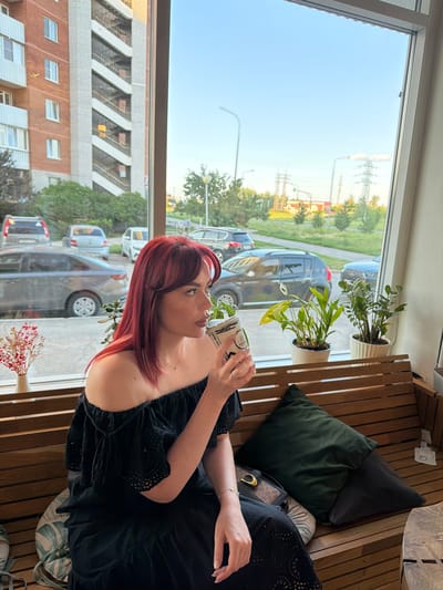 A woman with red hair sits on a wooden bench in front of a window. She is wearing a black off-the-shoulder dress and holding a cup to her lips. Outside the window, several cars are parked, and buildings and power lines are visible in the distance. The scene appears to be in the city of Timofeyevka, Russia.