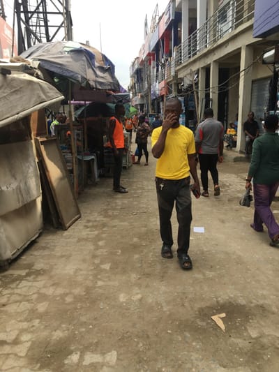 The image captures a busy market street scene in Ojo, Nigeria, under an overcast sky. The unpaved ground consists of uneven dirt and worn flagstones, with a small piece of brown cardboard and white paper visible as debris.
On the left side of the street, a series of makeshift market stalls are constructed with various tarpaulin and fabric coverings, supported by wooden frames. A large, dark metallic utility structure rises on the far left. On the right side, multiple multi-story commercial buildings, featuring balconies and exposed electrical wiring, line the street.
In the foreground, a male individual of apparent Nigerian descent walks towards the viewer. He wears a bright yellow short-sleeved t-shirt, dark trousers, and black sandals. His right hand is raised, touching his nose and mouth area. A set of keys is clipped to his left hip, and he holds a dark-colored object, possibly a mobile phone, in his left hand.
Further into the scene, on the left, another male subject in an orange sleeveless shirt stands near a market stall. In the midground, a male wearing a grey and red short-sleeved shirt and black trousers walks away from the camera, followed by a female in a dark green top, purple trousers, and carrying a black handbag. Numerous other individuals populate the background, engaged in various activities within the commercial street environment.