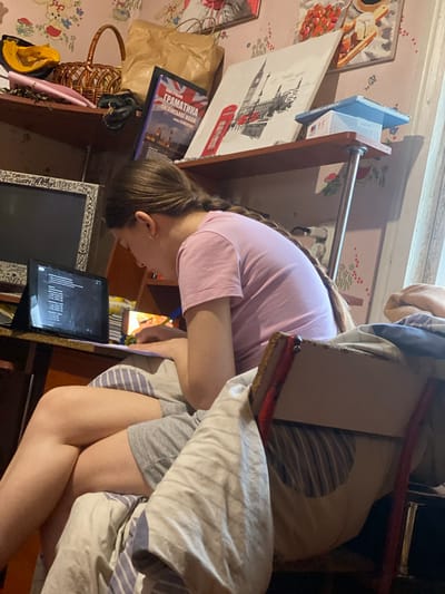 The image depicts a young woman with brown hair in a braid, wearing a pink t-shirt and grey shorts, sitting at a desk with her legs crossed. She is looking at a tablet device and appears to be studying or doing homework. The tablet displays a list of mathematical problems. She is also holding a pen and a sheet of paper.
The desk is cluttered with various items, including books, a basket, and a framed picture on the wall. The room appears to be a bedroom or study area, with wallpaper featuring a floral pattern. A window is visible in the background, and there is a chair with striped cushions. The lighting suggests it is daytime, possibly indoors with artificial light.