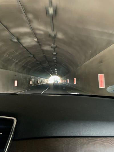 The image depicts a forward-facing view from inside a moving vehicle, traveling through a concrete road tunnel. The dashboard, dark in color with a textured finish, occupies the bottom quarter of the frame. A metallic-trimmed edge of an interior component is visible on the lower left. The tunnel, located in the vicinity of Бачковски манастир, Bulgaria, has an arched ceiling and light-colored concrete walls that show some darker discoloration. Overhead, linear light fixtures run along the ceiling. Along the right tunnel wall, several rectangular, vertically oriented red warning lights or reflectors are visible, spaced intermittently. A single white lane divider line is discernible on the asphalt road surface. Ahead, another light-colored vehicle, possibly a sedan or hatchback, is visible in the lane, approaching the brightly illuminated tunnel exit. The scene captures the vehicle's progression towards daylight at the end of the tunnel.
