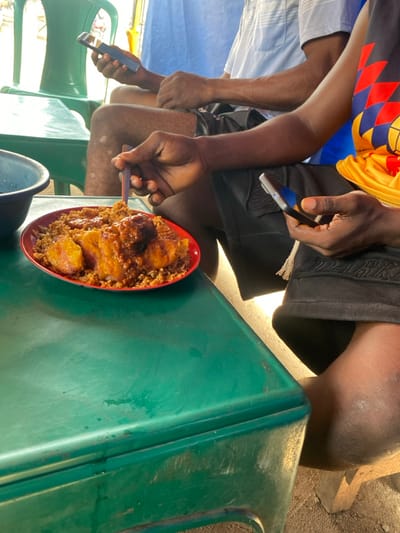 In Naze, Nigeria, two individuals are seated at a dark green, worn table in a casual outdoor setting. In the foreground, a person with dark skin wears black shorts and a partially visible colorful shirt. This person holds a dark smartphone in their left hand and a dark utensil in their right hand, poised over a red plate containing jollof rice, fried plantains, and a sauced item, possibly meat. A dark blue bowl is partially visible to the left on the table. In the background, a second individual, also with dark skin, wears a light blue collared shirt and black shorts, and is seated in a green plastic chair. This person holds a dark smartphone in their right hand. The ground is visible beneath the table, and a blue fabric element is present in the far background.