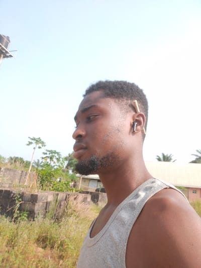 A young adult male, of dark skin tone, with short dark curly hair and a short beard, is depicted in profile, facing left, outdoors in Ishieke, Nigeria. He wears a light grey tank top. A dark-colored earbud is present in his right ear, with a light-colored wooden stick tucked behind it. The background features a clear, light blue sky, suggesting bright daylight conditions. To the far upper left, a metal structure supports a dark water tank. Below the horizon, a series of stacked concrete block structures are partially visible, surrounded by overgrown dry grasses and various green plants. Further in the background, a building with a light-colored roof is visible, partially obscured by trees, including several palm trees.