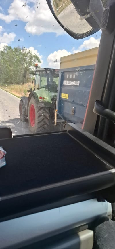 The media file shows a view from inside a vehicle, likely a tractor cabin, looking outwards through the windshield. The scene is outdoors on a sunny day, with a bright blue sky filled with white clouds.
The primary subjects are a green tractor and a large agricultural trailer. The tractor is positioned behind the trailer, with its front visible. The trailer is dark blue and has a yellow sticker on its side that reads "DO NOT transport damaged." There is also a license plate or identification number on the trailer which reads "+48 880 003 907". The tractor has large tires with red rims.
The setting appears to be a rural area, possibly a road or field in Bulgaria, given the location context provided. The ground visible is paved, suggesting a road. Trees and vegetation are visible in the background, further indicating an outdoor, rural environment. The overall mood is one of agricultural activity, with the equipment likely being used for farming tasks. The time of day is likely daytime, given the bright sunlight and clear sky. There are no people visible in the frame. The interior of the vehicle shows parts of the cabin, including a mirror and a dark surface that could be a dashboard or console. The windshield is dusty, with some insects visible on it.