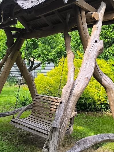 The image shows a rustic wooden swing set in a garden. The swing set is constructed from natural, unfinished logs, with a wooden seat suspended by chains. The roof of the swing set is made of wood shingles. In the background, there is lush green grass, bright yellow bushes, and a grey metal fence. Trees with green foliage are also visible. The overall scene suggests an outdoor recreational area. The location is identified as Žiaukos, Lithuania. There are no people in the image.