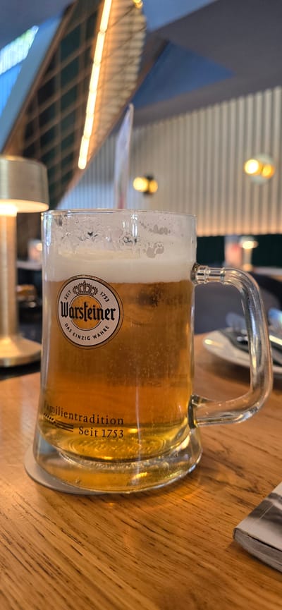 A close-up, eye-level shot focuses on a clear glass mug filled with golden-colored beer, topped with a frothy white head. The mug is positioned on a wooden table, with the handle facing the right side of the frame. Etched into the glass is a circular logo featuring a crown and the brand name "Warsteiner," along with the text "Seit 1753" and "Das einzig wahre Familien Tradition." The background is softly blurred, revealing hints of a restaurant or bar interior. To the left, a golden lamp is partially visible. In the background, there are vertical ribbed walls and some illuminated ceiling fixtures, suggesting an indoor setting. The lighting is warm and inviting. No individuals are present in the frame. The scene is a still life composition of a beverage. The image appears to be taken at the Warsteiner Brewery in Warstein, Germany, which is a well-known brewery and tourist destination. However, it is not in the city of Gütersloh, Germany.