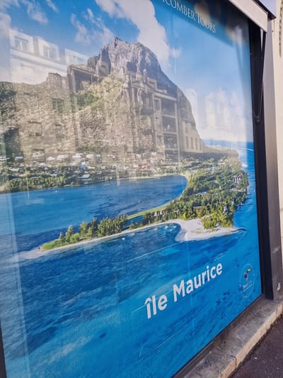 The image shows a large advertisement on the exterior of a building, likely a shop window or a storefront in Antony, France. The advertisement features a vibrant image of a tropical island with clear blue water and lush green vegetation, dominated by a large, rocky mountain in the background. Text overlays on the image read "Île Maurice" and "COMBER TOURS." There is also a logo for "TourCom" with a small bird graphic. The advertisement is reflecting the environment around it, including a building with windows and a cloudy blue sky. The bottom of the advertisement is bordered by a grey concrete curb and a dark pavement. The overall scene depicts an urban environment with commercial signage.