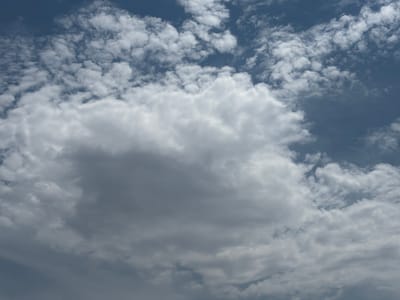 A close-up, eye-level shot captures a dramatic expanse of clouds against a deep blue sky. The clouds are varied in texture and density, with a large, dominant cumulus cloud taking center stage in the lower half of the frame. This central cloud is a mix of bright white highlights and darker, shadowed areas, suggesting it is thick and possibly casting its own shadow. Around it, smaller, wispy clouds are scattered across the sky, some appearing as delicate brushstrokes, others as more defined puffs. The sky itself transitions from a rich, dark blue at the top to a lighter, almost hazy blue towards the bottom, hinting at the cloud cover's effect on light diffusion. The overall impression is one of a dynamic and slightly overcast day, with the clouds appearing to be in motion. There are no people, objects, or discernible location cues in the image beyond the sky and clouds. The lighting suggests it is daytime, likely mid-afternoon given the intensity of the light on the clouds. The weather appears to be partly cloudy with potential for more significant cloud build-up. No text is visible.