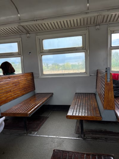 The image shows the interior of a train carriage with wooden seating. In the foreground, two wooden benches are visible. The bench on the left is positioned facing forward, while the bench on the right is positioned facing backward. Both benches are made of slatted wooden planks. A person with dark brown hair is seated on the left bench, facing away from the camera and looking out of the window. The windows of the train carriage offer a view of a rural landscape with trees and dry vegetation. The carriage appears to be old, with a simple interior design. The floor is a textured gray material. In the background, other seating arrangements and luggage are partially visible. The overall impression is of a functional, utilitarian train carriage.