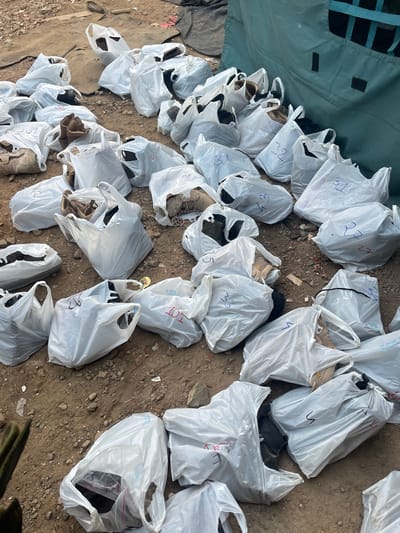 The image shows numerous white plastic bags scattered on a dirt and rock-strewn ground. Many of the bags are marked with numbers. Most bags appear to contain shoes, as glimpses of different shoe styles and colors are visible within the bags. The ground is uneven and appears to be outdoors. A section of green fabric, possibly part of a tent or tarp, is visible in the upper left corner. The location, Mira-Bhayander, India, is not discernible from the image.