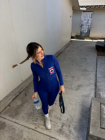 A woman in a blue, form-fitting suit stands on a concrete walkway. She wears white sneakers and has a long braid. She holds a water bottle in her left hand and a black face mask in her right. The suit has a "Cruz Azul" logo. The setting appears to be an alleyway with a white wall on one side and a concrete wall on the other, with a weathered door visible in the background.