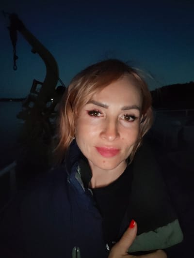 A medium-shot, outdoor, selfie-style photograph shows a woman with light brown hair and fair skin, wearing a dark jacket over a black shirt. She has on pink lipstick and pink eyeshadow. Her right hand is visible, with red nail polish on her thumb. The background is dark, with a dark blue sky and silhouetted trees on the right. A large, dark object with a curved arm and a hook is visible on the left side of the frame. The overall lighting suggests it is dusk or nighttime. The location is not discernable from the image.