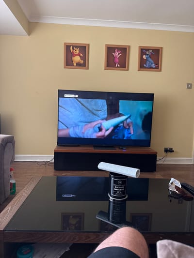 A living room scene is depicted with light yellow walls adorned with three Winnie the Pooh character pictures. A television is mounted on the wall above a brown wood cabinet, displaying a video of a woman in a blue shirt. To the right, a black glass coffee table holds a roll of paper towels, a candle labeled "Black Rose Sandalwood," and some debris. Parts of a person and a grey dog bed are visible at the bottom of the image. The location is Colchester, United Kingdom.