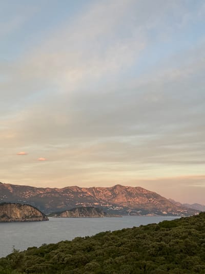 A scenic view of a coastal mountain range overlooking a body of water at dusk, likely near Višnjevo, Montenegro. The sky is a gradient of soft blues and pinks, with wispy clouds scattered throughout. In the foreground, a lush, dark green forested hillside slopes down towards the water. The water itself is a deep, calm blue, reflecting the muted colors of the sky. Across the water, a rugged mountain range rises, its peaks illuminated by the warm, fading sunlight, casting long shadows. Small, white structures, possibly buildings, are visible nestled along the slopes of the mountains. Two rocky islands, one larger and more prominent than the other, rise out of the water in the mid-ground, their surfaces textured and weathered. The overall atmosphere is serene and tranquil.