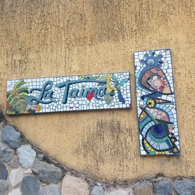 The image captures a section of a textured wall adorned with two rectangular mosaic art pieces and a stone foundation below. The wall itself is a light tan or yellowish-brown color with a rough, stucco-like finish, showing some darker vertical streaks.
The main mosaic on the left spells out "La Taina" in dark blue-green, curved mosaic tiles. The letters are set against a background of small white mosaic squares. The artwork is embellished with natural motifs: green leaves and yellow flowers are integrated into the "L" and "a" of "La", and a small red heart is visible below the "i" in "Taina". A stylized hummingbird, also made of mosaic tiles in blue, green, and yellow, is depicted near the end of the word.
To the right, a taller, narrower mosaic piece presents an abstract, colorful design. It features various shapes and patterns in shades of blue, green, brown, pink, yellow, and black, all outlined with dark grout lines against a white mosaic background. The design includes circular and curvilinear elements, possibly representing stylized birds or organic forms.
Below the textured wall, a foundation made of irregularly shaped, multi-colored stones is visible, mortared together. The lighting suggests it is daytime, with bright, even illumination. The scene depicts decorative elements on an exterior wall in El Limon, Venezuela.