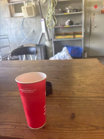 The image shows a red paper cup with text on it, resting on a wooden surface. The cup has a black lid. The text on the cup reads "EurothermenResort BAD SCHALLERBACH". In the background, shelves filled with various items, including a blue plastic bin and some white material, are visible. There is also a glimpse of what appears to be a chair and some electrical wiring. The scene suggests an indoor setting, possibly a workshop or storage area. The lighting appears to be artificial, indicating it could be any time of day. The overall impression is one of a casual, everyday environment.