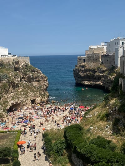 This image captures a vibrant, sunny day at a crowded beach and cove nestled among cliffs in Polignano a Mare, Italy. The scene is dominated by a narrow, sandy beach, teeming with numerous people. Many individuals are seen sunbathing on towels spread across the sand, while others seek shade under a scattering of colorful beach umbrellas in shades of red, blue, yellow, and white. Adjacent to the beach, a clear, turquoise-blue sea fills a small cove, where many people are swimming, wading, and enjoying the water.
The beach and cove are dramatically framed by towering, rugged rocky cliffs. Perched atop these cliffs, particularly on the left side and extending across the upper edge of the frame, are numerous white-washed buildings with distinctive Mediterranean architecture, indicating a historic town overlooking the sea. Some groups of people are visible near these buildings, possibly on viewpoints. On the right side of the image, lush green bushes and trees grow along the upper edges of the cliffs, adding a touch of natural verdancy. A paved pathway is visible in the foreground on the right, with a few people walking along it.
The clear, brilliant blue sky suggests excellent weather, and the strong, direct sunlight indicates it is likely midday or early afternoon. The overall atmosphere is one of bustling summer recreation and enjoyment, characteristic of a popular tourist destination. No discernible text is visible in the image.