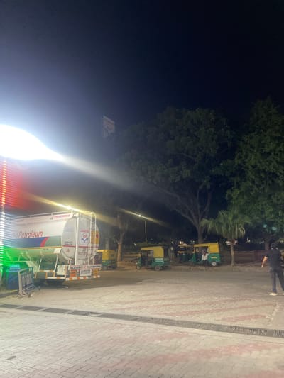 The image depicts a night scene at a fuel station in Kajheri, India. A large tanker truck, branded "Pakistan Petroleum" and "POWER PROGRESS," is parked, its side illuminated by bright lights, creating lens flares. The tanker displays HP logos and capacity information. Several auto-rickshaws, typical of Indian city transport, are lined up on the side of the road. One auto-rickshaw has passengers inside. A person is standing near a fuel pump. In the background, lush green trees are visible against the dark night sky. The ground is paved with interlocking concrete blocks and features a drainage grate. A "NO PARKING" sign is also present in the foreground.