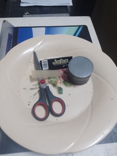 The image displays an overhead view of a light-colored, round plate, possibly beige, bearing a faint floral pattern, situated on a lighter, flat surface. The plate partially obscures a blue and white patterned item beneath it. On the plate, a pair of black-handled scissors with red finger loops and partially open silver blades rests. Adjacent to the scissors is a black packet of "Jaba ULTRA FINE PAPER No. 70" rolling papers. A single, light-beige rolling paper is laid flat on the plate, positioned beneath the scissors and the paper packet. To the right, a silver-colored, cylindrical herb grinder is present. Numerous small, dark green fragments, consistent with processed plant material, are dispersed across the plate's surface, alongside larger, unground pieces of similar vegetation. The background reveals stacked white papers and a small, irregularly shaped golden object. The arrangement of objects indicates activities associated with the preparation of plant material. This scene is located in Ziwa La Ng'ombe ward, Kenya.