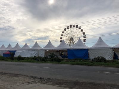 The image depicts an outdoor scene in Blangbitra, Indonesia, featuring a temporary event setup under an overcast sky with bright sunlight breaking through. In the midground, a row of at least eight white, conical-roofed tents (pagoda tents) extends horizontally. A small red tent is partially visible behind the leftmost white tent, and one of the tents further right has a blue tarp serving as a wall. Behind these tents, a large Ferris wheel with multiple passenger gondolas is prominently displayed, its intricate metal structure visible against the sky. Multiple power lines traverse horizontally across the upper midground. The foreground consists of an asphalt road, likely two lanes, running horizontally across the lower third of the frame, with a faint yellow line visible on the right side. A strip of unkempt grass and sparse shrubs separates the road from the tents. No individuals are visible in the scene.