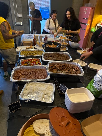 The image shows a food buffet setup, likely for a gathering. The buffet includes multiple aluminum trays containing various foods such as shredded beef, carne asada, beans, rice, sauteed peppers and onions, and corn. Next to the food trays are stacks of tortillas and bowls. People are shown serving themselves from the buffet. The location is Culver City, United States of America based on a recycling bin in the background.