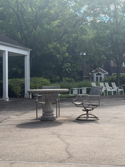 This image depicts an outdoor patio area in Edison, New Jersey, United States. The scene includes a concrete patio surface, a white stone table with decorative trim and metal legs, and a metal patio chair with a swivel base. In the background, there are several white lounge chairs, a glimpse of a building with dormer windows, and dense foliage from trees and shrubs. A white porch or patio structure with supporting columns is also visible. The lighting suggests it is daytime.