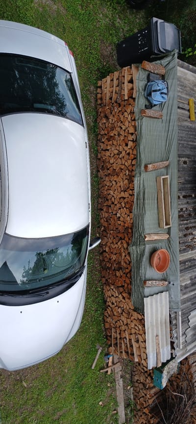 In an outdoor setting, likely a yard in Võhma, Estonia, a white car is parked on the left side of the image. To the right of the car, a large stack of chopped firewood is neatly piled. Beyond the firewood, a green tarp covers a surface, upon which several items are placed: a blue backpack, two pieces of log, a terracotta pot, and a wooden rectangular frame. Further to the right, what appears to be a makeshift roof structure made of corrugated panels is visible. A black trash bin is situated behind the firewood stack. On the ground near the car and the firewood, two axes and a smaller blue object, possibly another tool or a piece of equipment, are present. The ground is covered in grass and some dirt. The overall scene suggests preparation for a task involving firewood, possibly for heating or a similar purpose.