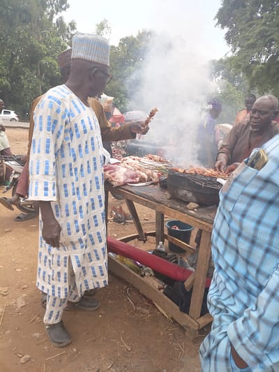 In the city of Tilde, Nigeria, a street vendor is grilling meat skewers over a charcoal grill. The vendor, an older man with glasses and a patterned cap, is wearing a blue and white patterned robe. He is standing next to a wooden table laden with raw meat. Smoke billows from the grill, obscuring some of the background details. In the foreground, a person in a blue checkered shirt is visible. The ground is dirt, and trees and vegetation are in the background. Other individuals are present, some appearing to be customers or associates of the vendor. A white car is visible in the distance on a road.