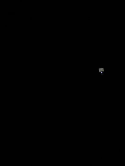 The image depicts an extremely dark environment, primarily black, suggesting a nocturnal scene or severe low-light conditions in Easingwold, United Kingdom. In the upper-right quadrant of the frame, a small, rectangular, illuminated object is visible. This object appears to be a sign or a digital display, exhibiting indistinct text or a graphic on a light-colored background. Directly beneath this illuminated object, a distinct, faint blue light source is present. No people, specific actions, or other discernible subjects or objects are visible due to the prevailing darkness. The environment is devoid of detail beyond these two light sources.