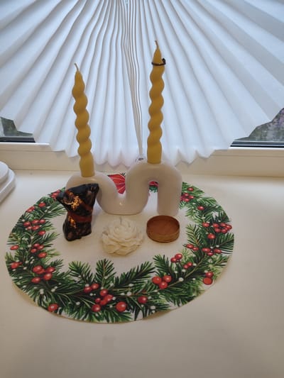 The image displays a decorative arrangement on a white surface, likely a windowsill, in front of a white pleated blind. Two twisted, yellow candles are held upright by a white, wavy candelabra. On a round placemat adorned with a festive Christmas wreath pattern of evergreen branches and red berries, are three small objects: a dark, possibly ceramic, figurine of a dog with a red and gold collar, a white rose-shaped candle, and a small wooden bowl. The scene appears to be indoors, with natural light filtering through the window, suggesting it might be daytime. The overall impression is one of seasonal decoration, possibly for the holidays. No text is visible in the image. The location context of Lisakovsk, Kazakhstan is noted.